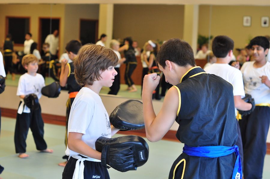 One young martial arts student holds padded mitts for another student to practice strikes. Behind them, you can see a full class in a mirrored wall.