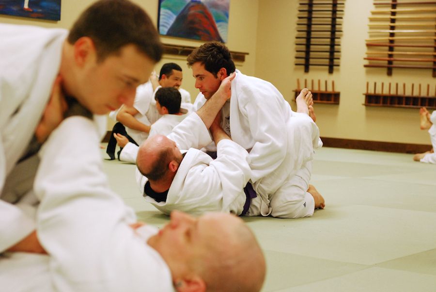 In a clean & well-lit dojo on tatami mats, 6 students are on the ground in 3 pairs practicing Brazilian Jiu-Jitsu in white gis. On the walls of the dojo behind them are a painting of a man meditating atop a hill, and a rack of wooden weapons.