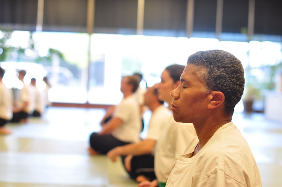 Class of students meditating in a dojo as a lesson in mindfulness. In the foreground, a woman sits in profile, eyes closed.