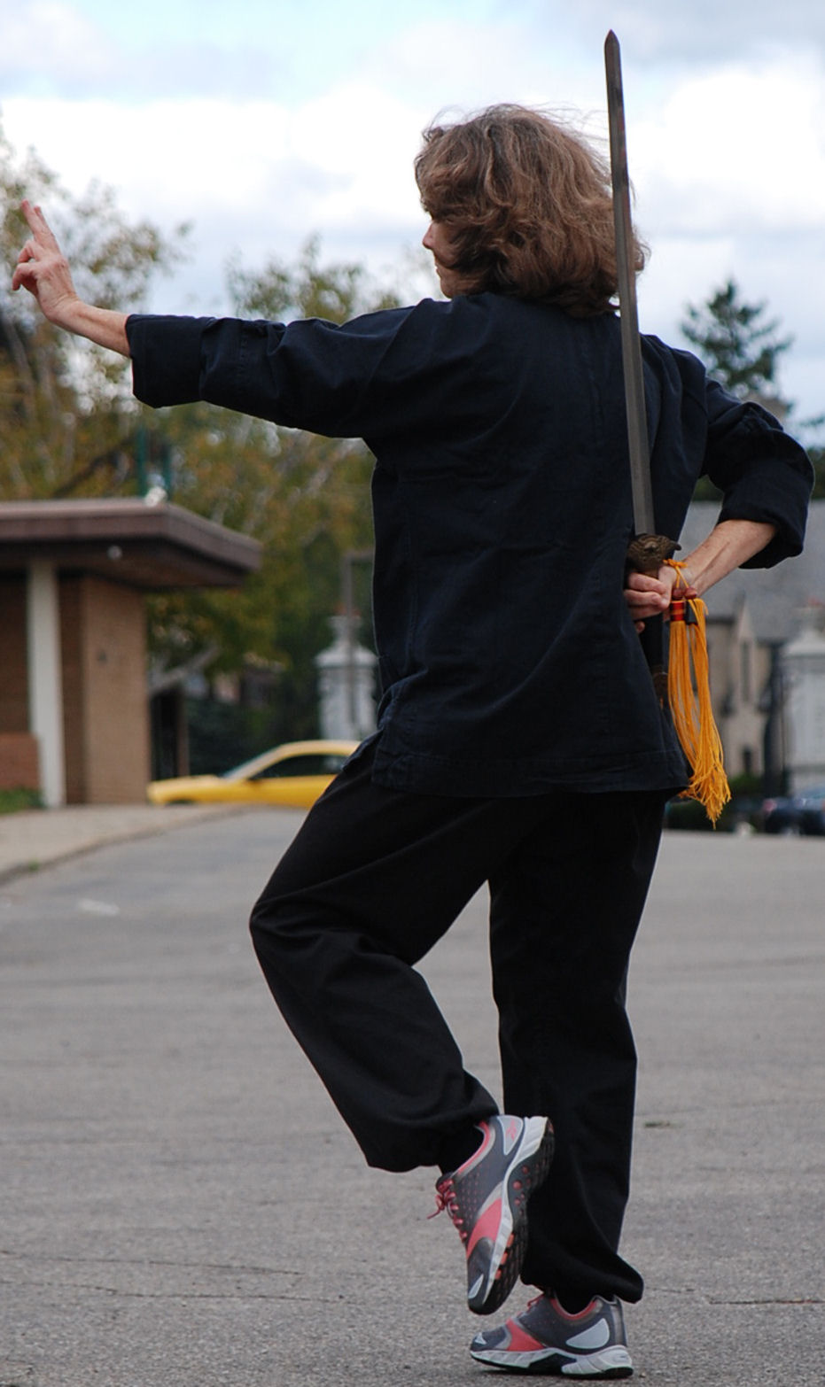 A woman holds a tai chi sword behind her back with her other hand outstretched, palm out, and one foot slightly lifted with toes pointed down.