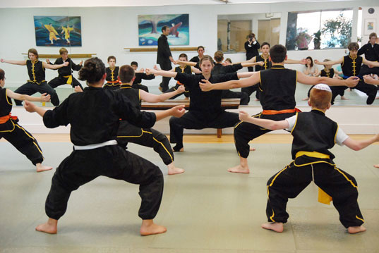 An adult woman leads a class of young martial arts students in holding a "horse stance" pose.
