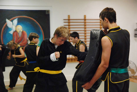Teenagers in martial arts uniforms practice strikes on large pads held by another student.